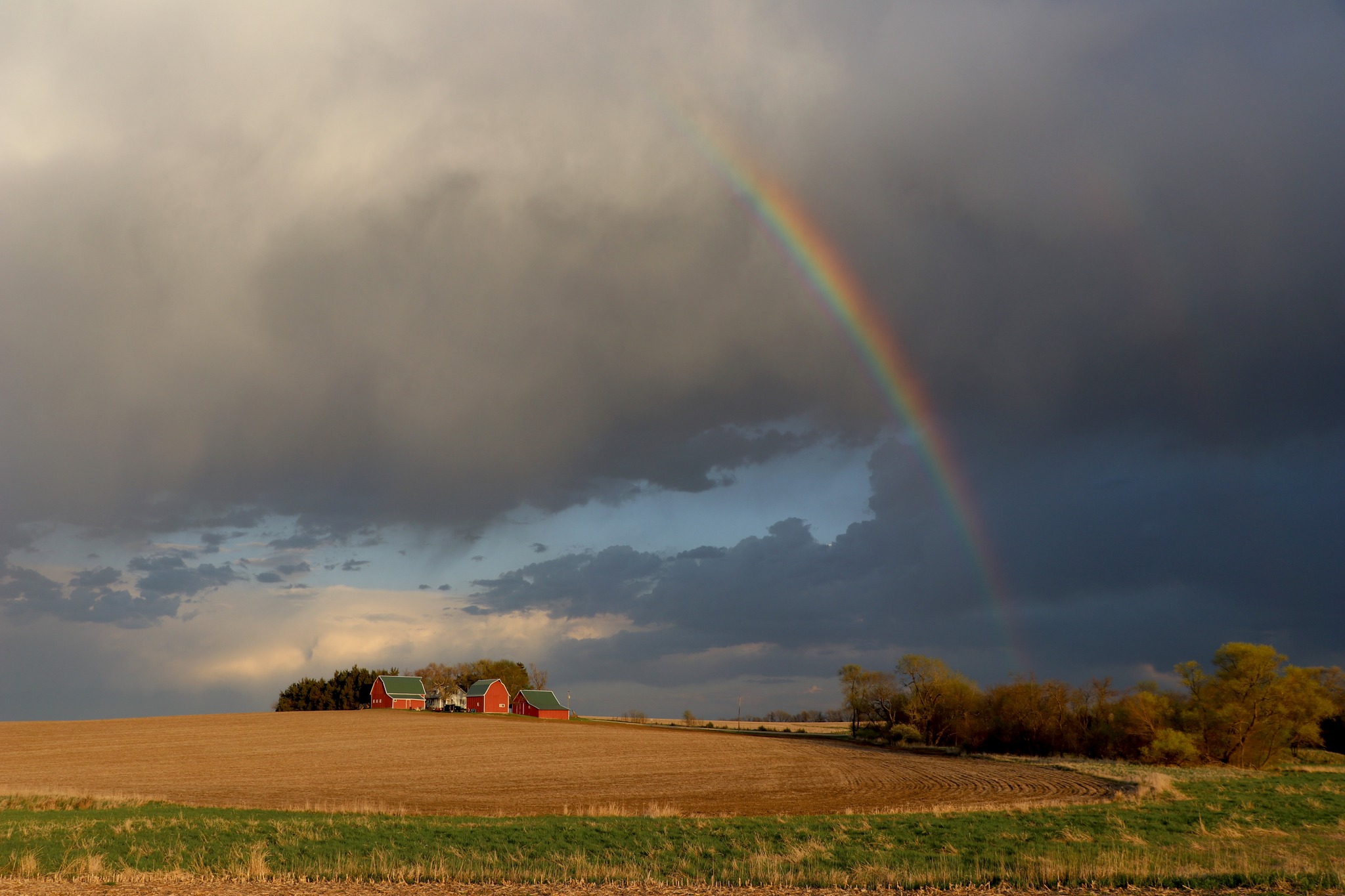 A farmstead below dark clouds and a rainbos.